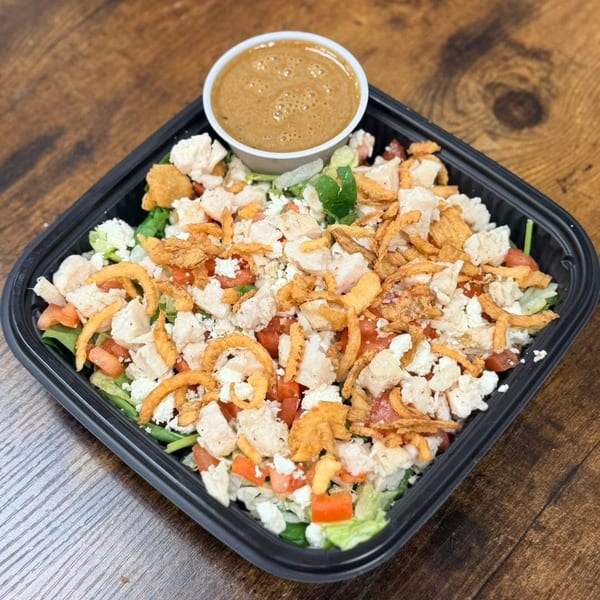 A salad with chicken, tomatoes, feta, crispy onions, and dressing in a black container on wood.
