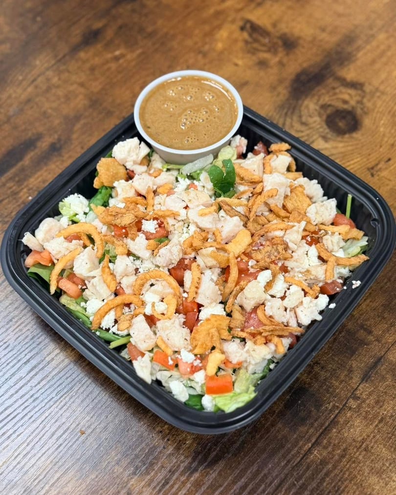 A salad with chicken, tomatoes, feta, crispy onions, and dressing in a black container on wood.