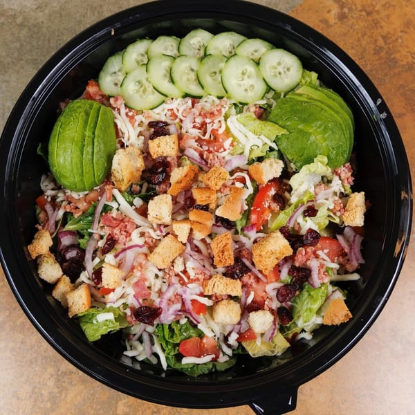 A vibrant salad in a black bowl, topped with avocado, cucumbers, croutons, cheese, and dried cranberries.