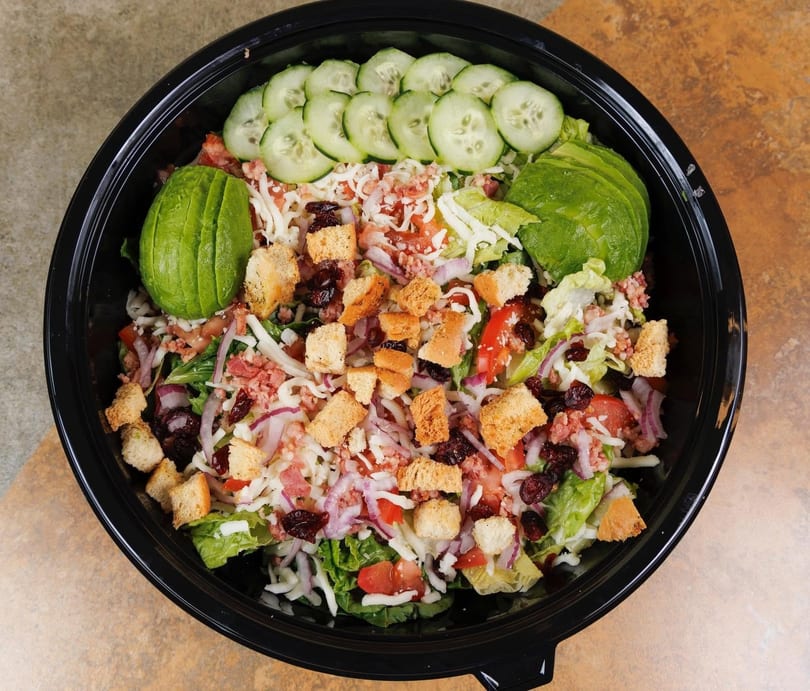 A vibrant salad in a black bowl, topped with avocado, cucumbers, croutons, cheese, and dried cranberries.