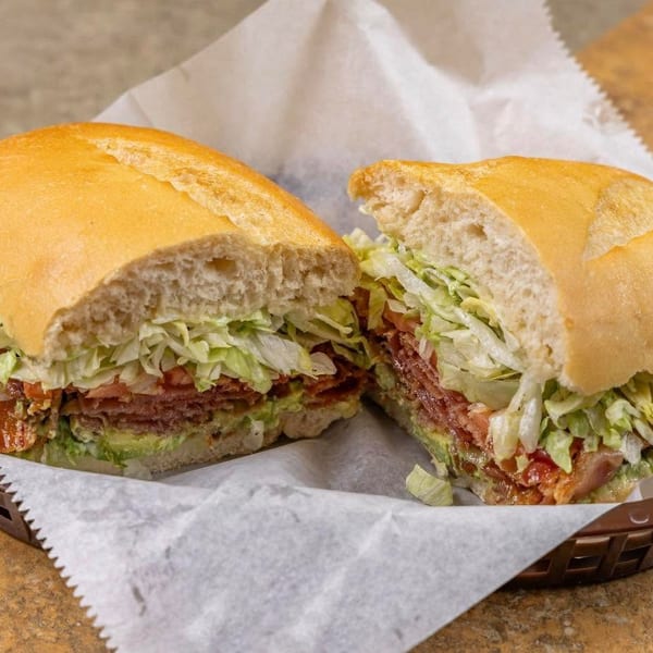 A large sandwich, cut in half, filled with meat, lettuce, tomato, and avocado in a basket.