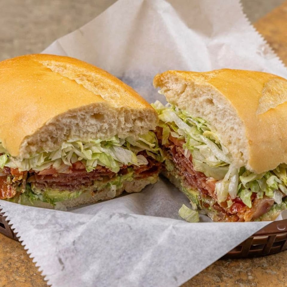 A large sandwich, cut in half, filled with meat, lettuce, tomato, and avocado in a basket.