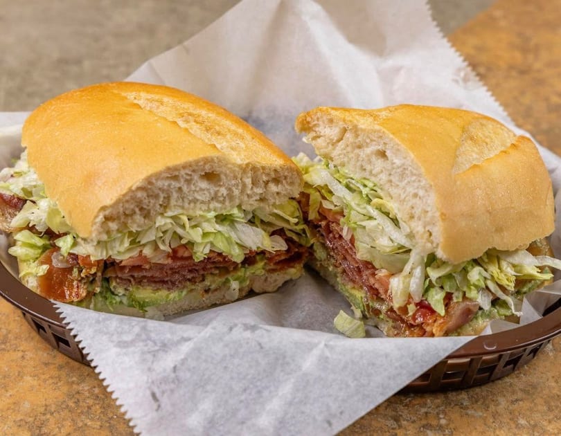 A large sandwich, cut in half, filled with meat, lettuce, tomato, and avocado in a basket.